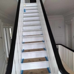 Magmax - Białe schody z czarną balustradą w trakcie zabezpieczania stopni niebieską taśmą malarską przed pracami wykończeniowymi.