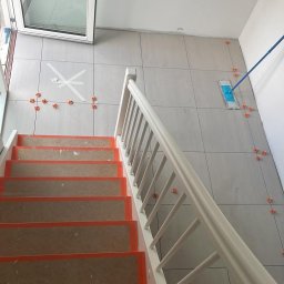 Magmax - Układanie płytek podłogowych na korytarzu z widocznymi krzyżykami dystansowymi i schodami zabezpieczonymi kartonem.