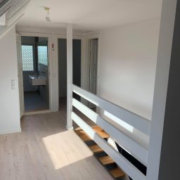 Magmax - Jasne wnętrze po remoncie: świeżo pomalowane ściany, jasna drewniana podłoga, biała balustrada schodów, widok na łazienkę z umywalką.