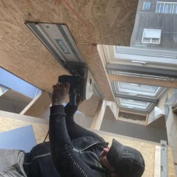 Magmax - Pracownik z tatuażami na rękach tnie płytę OSB pilarką szynową Makita, widoczne okna dachowe i poziomica w tle.