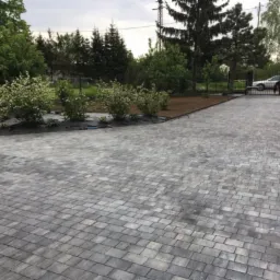 Ułożona szara kostka brukowa na podjeździe, z widocznymi krzewami i drzewami w tle. W oddali brama wjazdowa i samochód. Perspektywa z dołu podkreśla rozległość powierzchni.