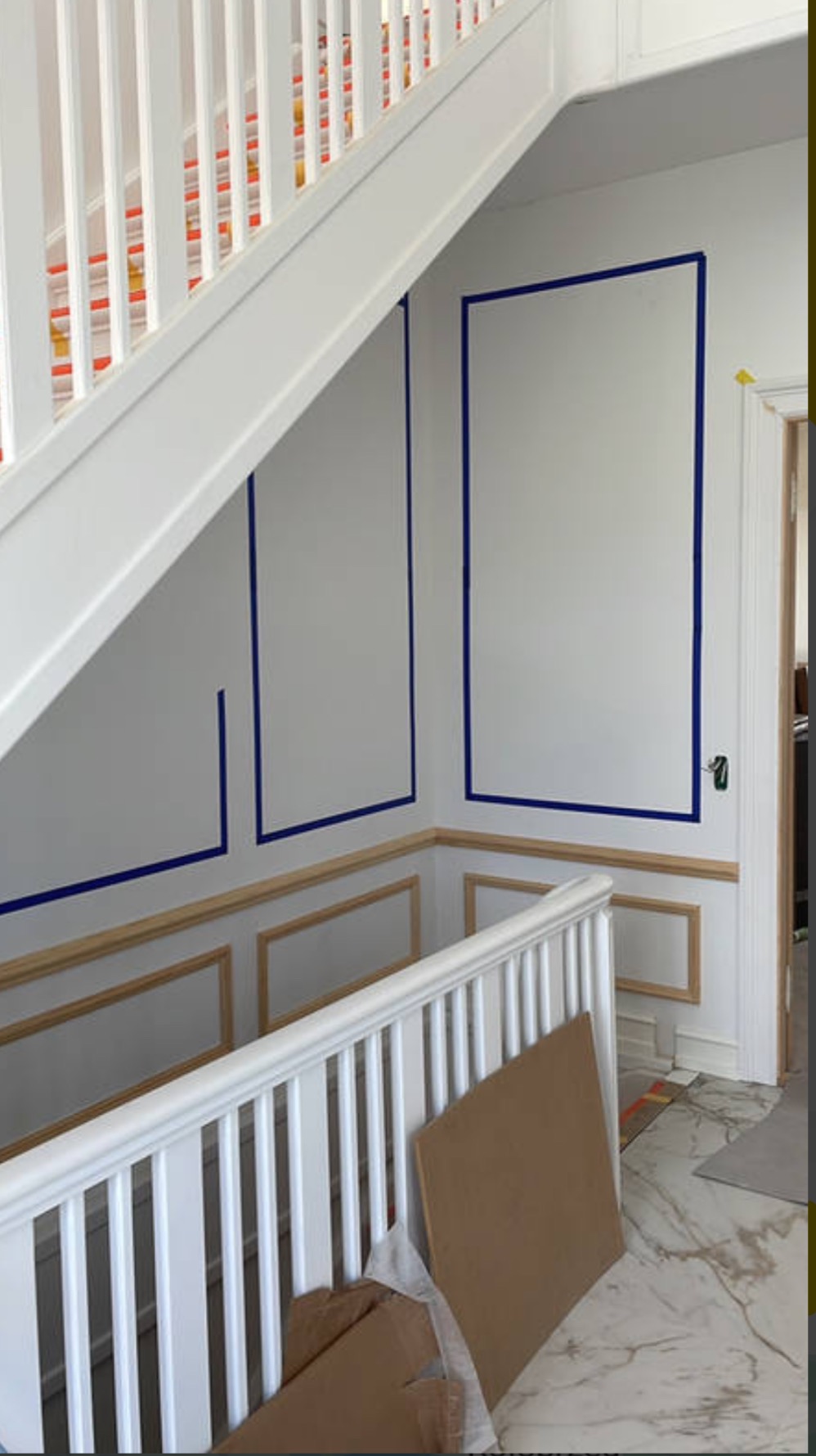 Klatka schodowa w trakcie prac wykończeniowych: ściany z białymi panelami obramowanymi niebieską taśmą malarską, drewniane listwy dekoracyjne, marmurowa podłoga, białe schody z kolorową wykładziną...