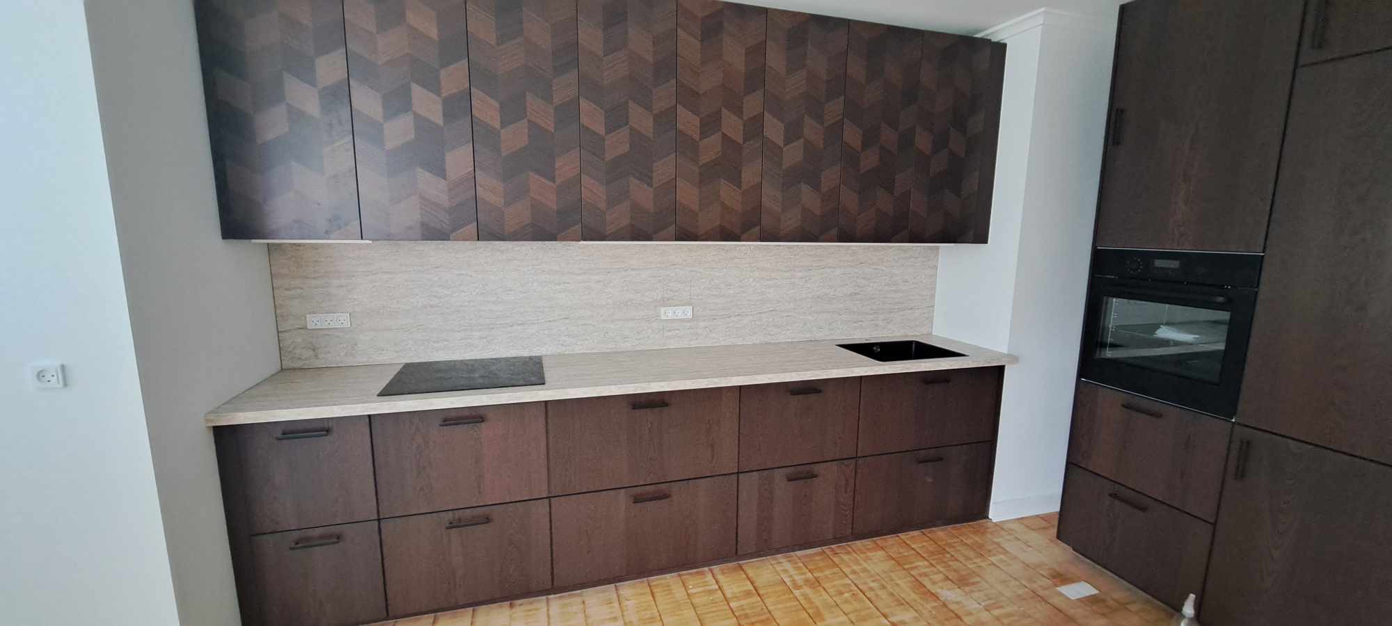 Nowoczesna kuchnia z ciemnobrązowymi szafkami o geometrycznym wzorze na górnych frontach, jasnym blatem i płytkami nad blatem, czarną płytą grzewczą i zlewem, oraz wbudowanym piekarnikiem.