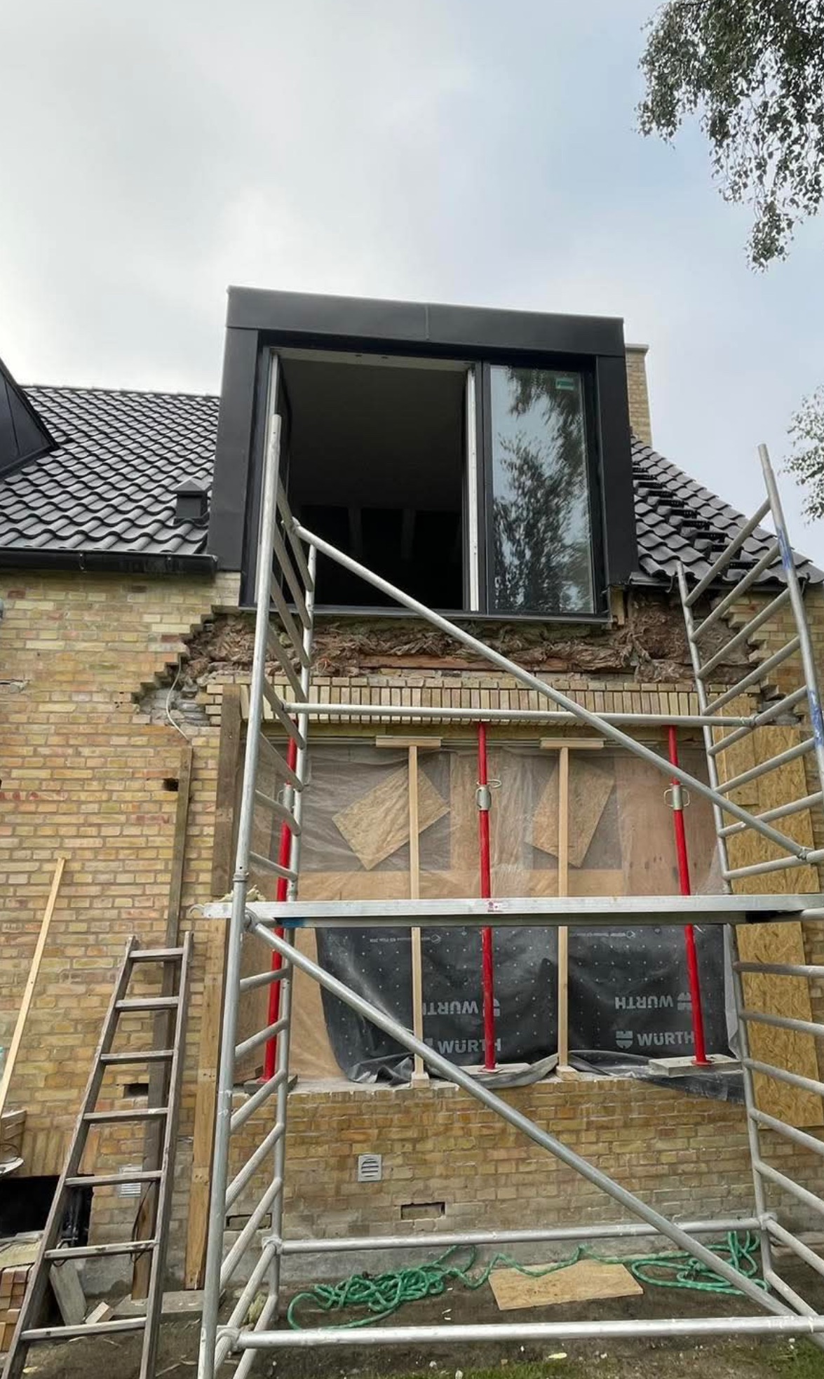 Dom z cegły z zamontowanym oknem dachowym, widoczne rusztowanie i uszkodzona ściana zewnętrzna w trakcie naprawy.