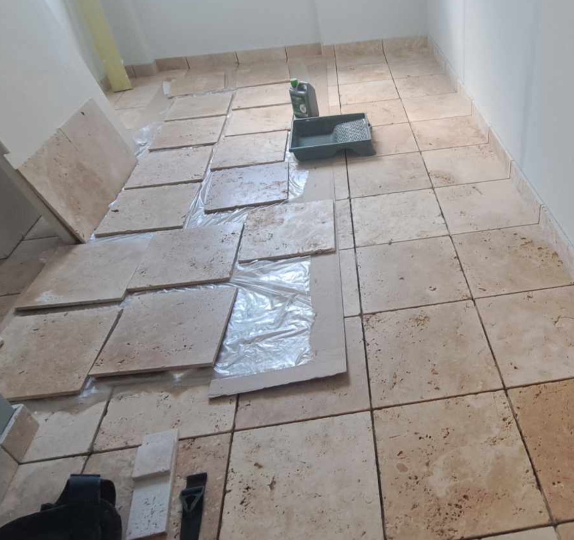 Kładzenie beżowych, kwadratowych płytek podłogowych z widocznym pojemnikiem na farbę i folią ochronną na części płytek.