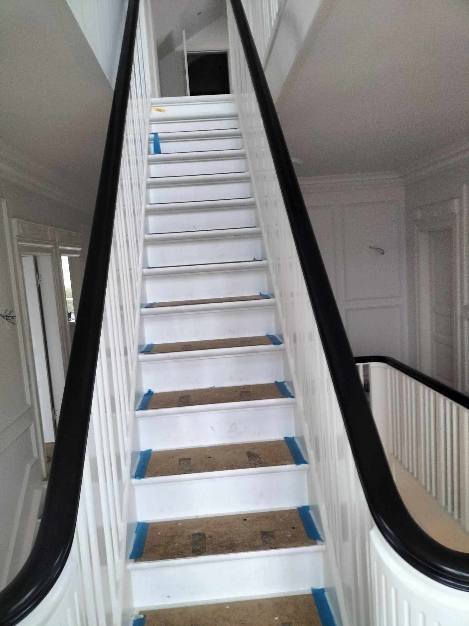 Białe schody z czarną balustradą w trakcie zabezpieczania stopni niebieską taśmą malarską przed pracami wykończeniowymi.