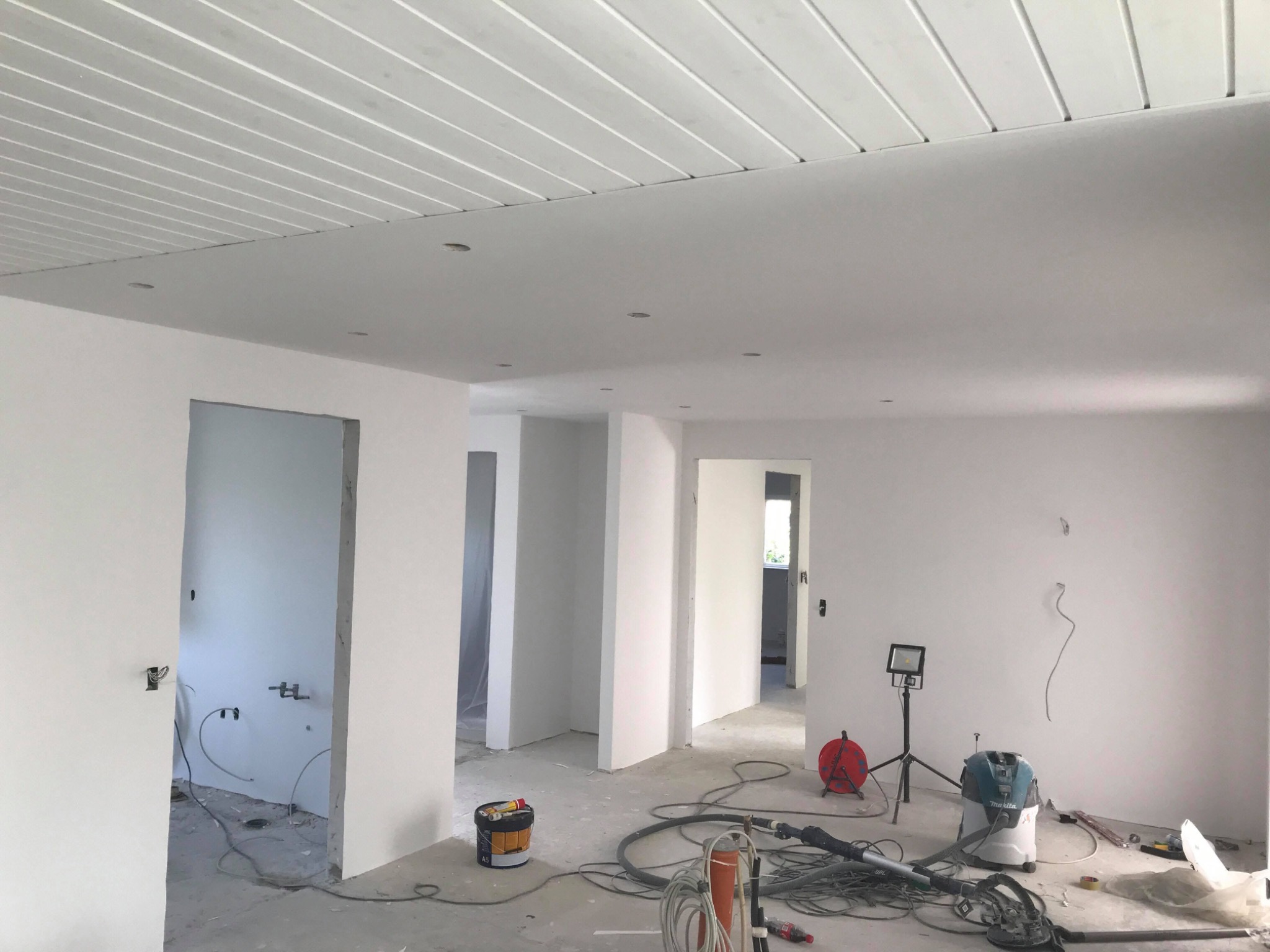 Wnętrze w trakcie remontu: białe ściany, sufit z paneli, widoczne narzędzia budowlane, kable i lampa oświetleniowa na trójnogu.