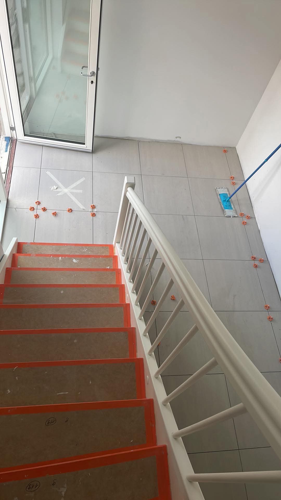 Układanie płytek podłogowych na korytarzu z widocznymi krzyżykami dystansowymi i schodami zabezpieczonymi kartonem.