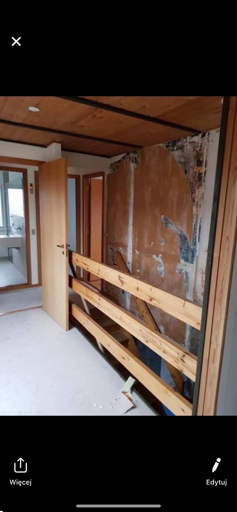Wnętrze w trakcie remontu: odsłonięta ściana z resztkami starej farby, drewniana konstrukcja zabezpieczająca, widok na łazienkę z białymi płytkami w tle.