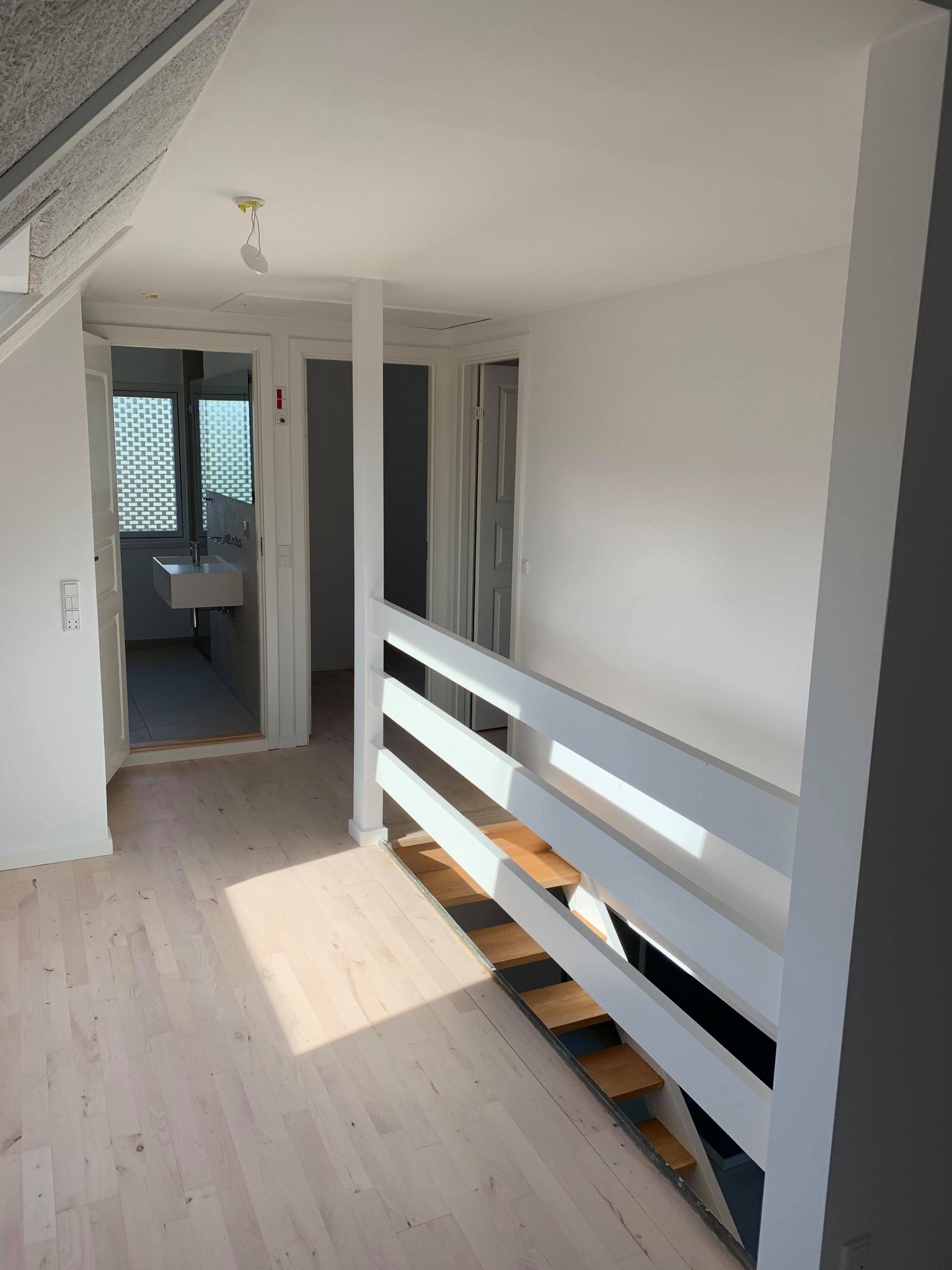 Jasne wnętrze po remoncie: świeżo pomalowane ściany, jasna drewniana podłoga, biała balustrada schodów, widok na łazienkę z umywalką.