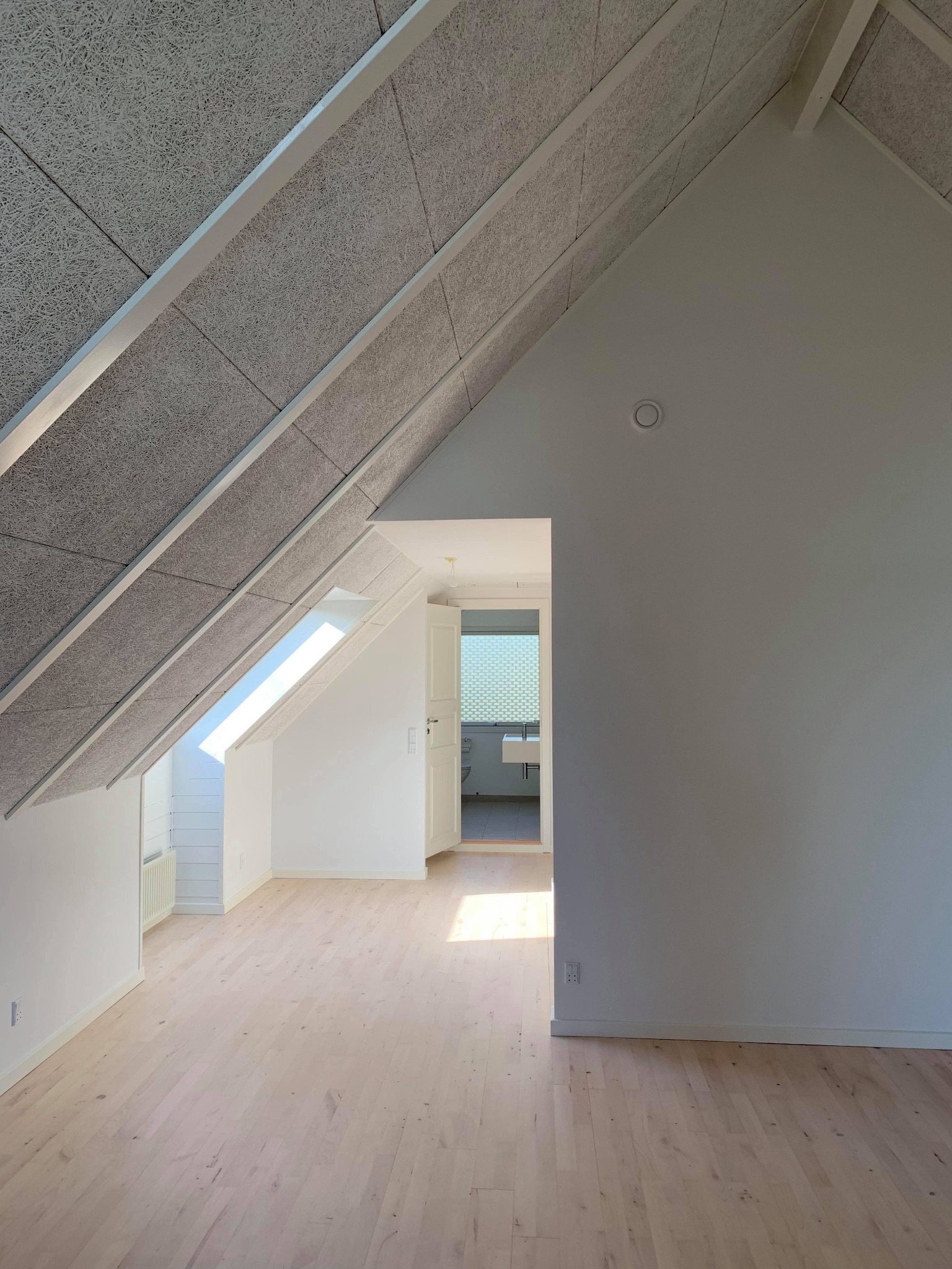 Poddasze z wykończonymi ścianami w jasnym odcieniu, drewnianą podłogą, oknem dachowym wpuszczającym światło i szarą, akustyczną płytą sufitową z widocznymi belkami stropowymi.