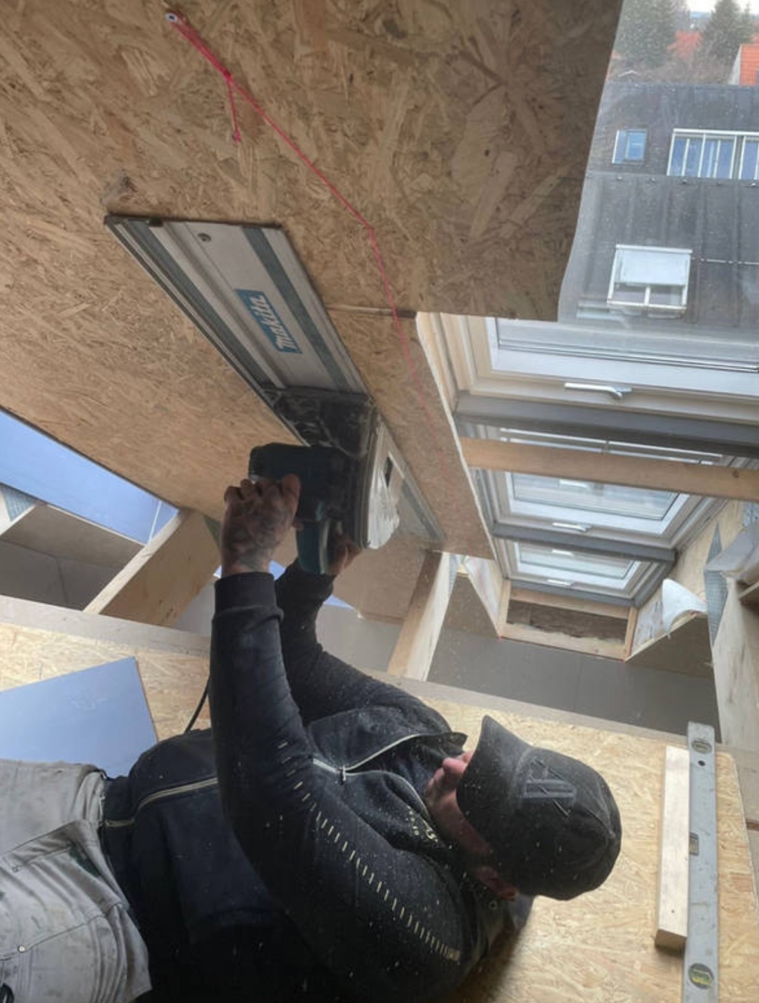 Pracownik z tatuażami na rękach tnie płytę OSB pilarką szynową Makita, widoczne okna dachowe i poziomica w tle.