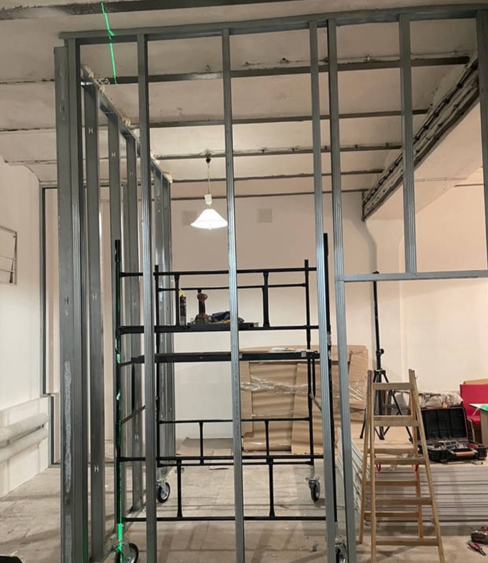 Montaż metalowej konstrukcji szkieletowej ścian działowych w trakcie prac wykończeniowych, widoczny laserowy niwelator, rusztowanie na kółkach i drabina.