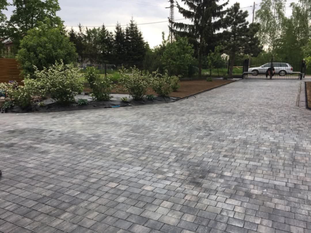 Ułożona szara kostka brukowa na podjeździe, z widocznymi krzewami i drzewami w tle. W oddali brama wjazdowa i samochód. Perspektywa z dołu podkreśla rozległość powierzchni.