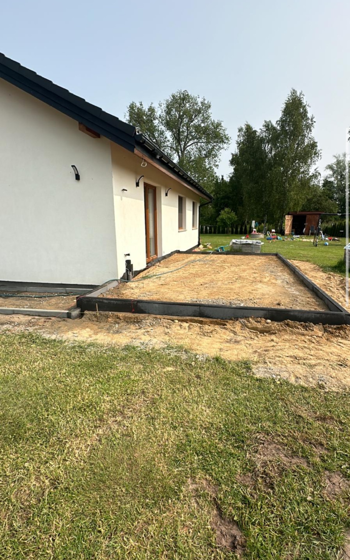 Przygotowanie podłoża pod taras z obrzeżami z kostki, widok na dom z ogrodem w tle. Etap prac ziemnych przed ułożeniem nawierzchni.