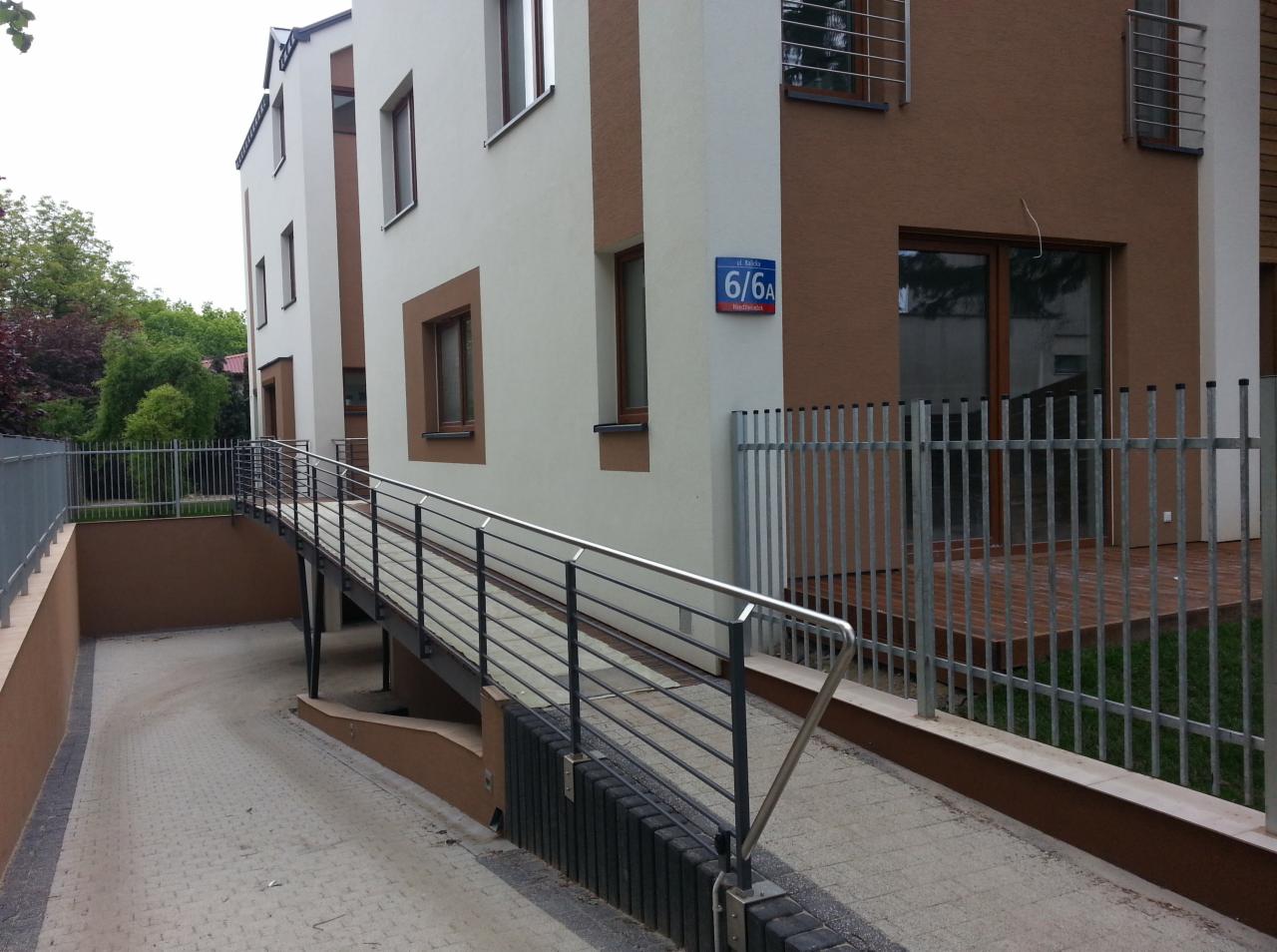 Nowoczesny budynek mieszkalny z podjazdem dla niepełnosprawnych, widoczny numer budynku 6/6A, metalowe ogrodzenie i drzewa w tle.