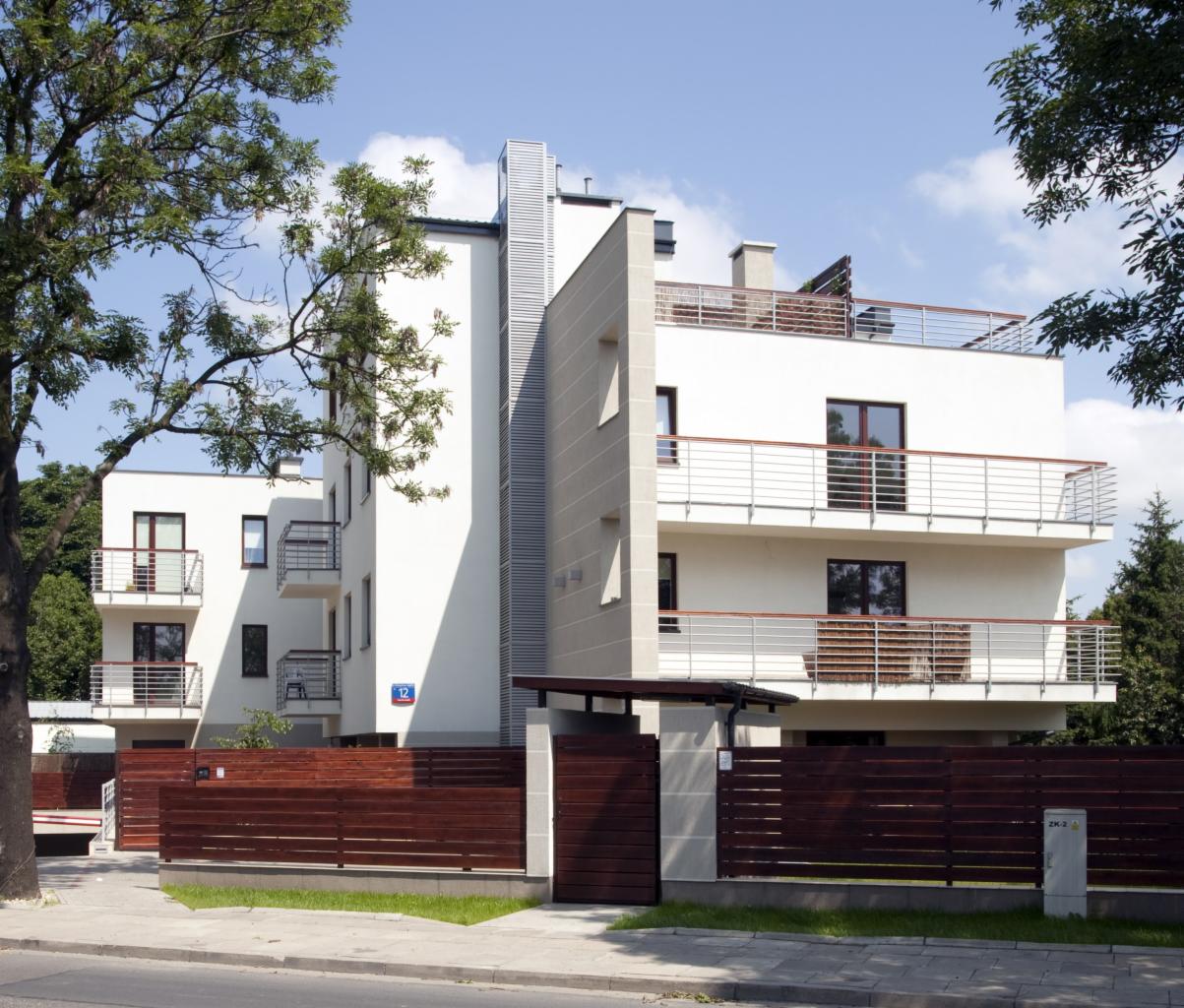 Nowoczesny budynek apartamentowy z balkonami i drewnianym ogrodzeniem, widoczny znak z numerem '12' na ścianie.