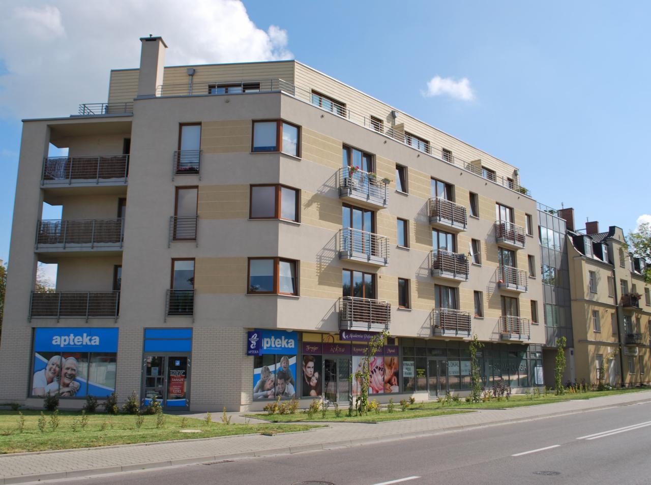 Nowoczesny budynek mieszkalny z balkonami i lokalami usługowymi na parterze, w tym apteki, sfotografowany w słoneczny dzień.