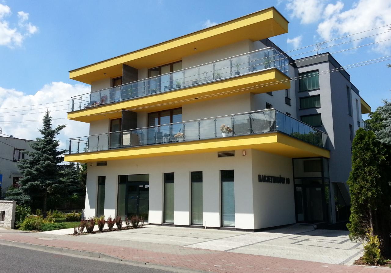 Trzypiętrowy budynek mieszkalny z żółtymi balkonami i szklanymi balustradami, widok z ulicy w słoneczny dzień.