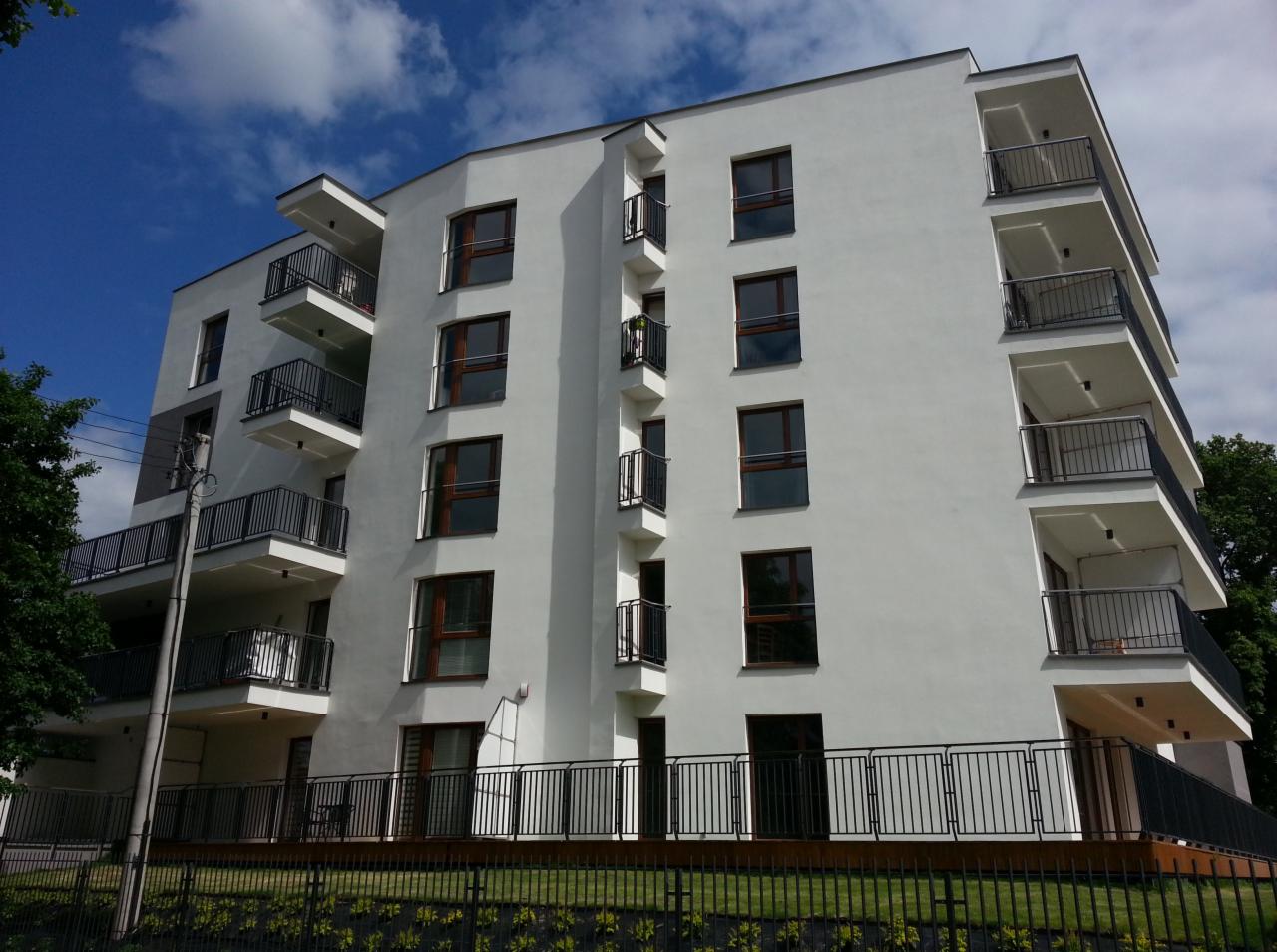 Nowoczesny budynek apartamentowy z białymi elewacjami, ciemnymi ramami okiennymi i balkonami z czarnymi balustradami, sfotografowany w słoneczny dzień z niskiej perspektywy.