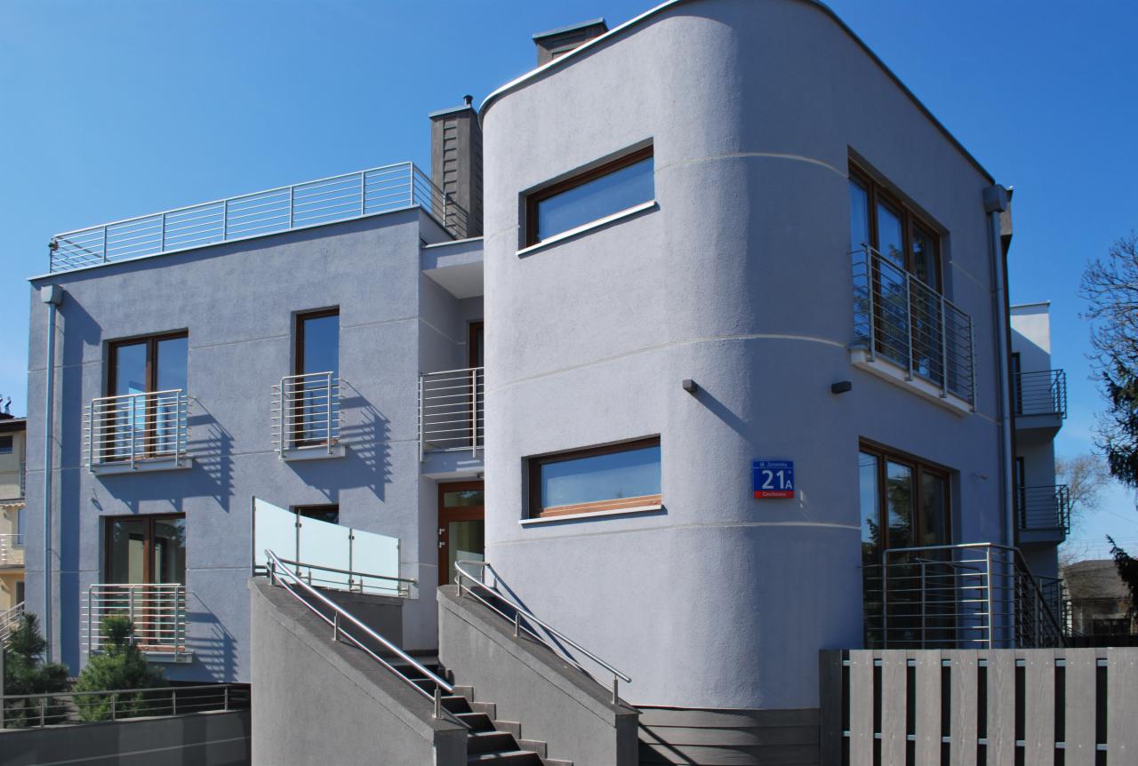 Nowoczesny, dwupiętrowy budynek mieszkalny z zaokrąglonym narożnikiem i balkonami ze stali nierdzewnej, widoczny znak z numerem '21A'.