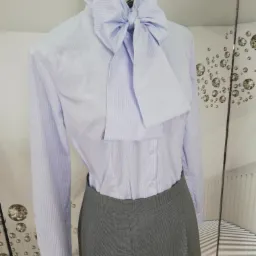 Jasnoniebieska koszula w prążki z dużym wiązaniem pod szyją, zestawiona z szarą spódnicą w drobną kratkę, prezentowana na manekinie.