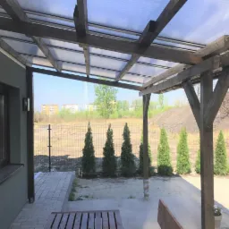 Zadaszony taras z drewnianą konstrukcją, widok na ogród z rzędem małych iglaków i budynki w oddali.