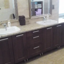 Łazienka z brązowymi szafkami na wymiar z granitowym blatem, podwójna umywalka, lustra w srebrnych ramach, sterta ręczników i pralka z przodu.