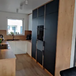 Nowoczesna kuchnia z białymi szafkami dolnymi i drewnianymi blatami, czarne szafki wiszące z piekarnikiem w zabudowie, drewniana podłoga, widok na okno i drzwi do garderoby.