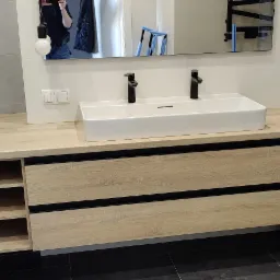 Nowoczesna łazienka z szafką pod umywalką z jasnego drewna z czarnymi akcentami, podwójna umywalka nablatowa i bateria w kolorze czarnym, na ścianie płytki w odcieniach szarości, w tle dekoracyjna...