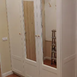 Biała szafa trzydrzwiowa z lustrami wbudowanymi w dwoje drzwi, z widocznym w lustrzanym odbiciu wnętrzem przedpokoju z wieszakami i drabinką.