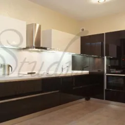 Nowoczesna kuchnia na wymiar z czarnymi, błyszczącymi frontami, białymi szafkami wiszącymi i stalowym okapem, oświetlona listwą LED pod szafkami.