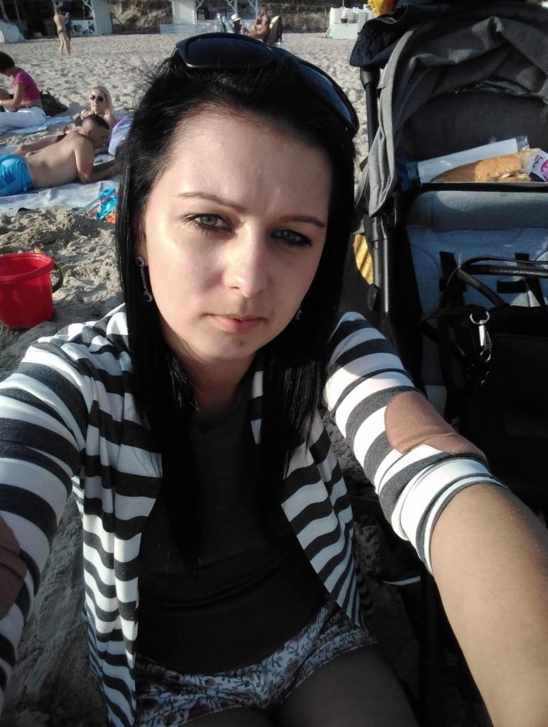 Osoba w paski kardigan siedzi na piasku obok wózka dziecięcego na plaży. W tle opalający się ludzie i budynki.