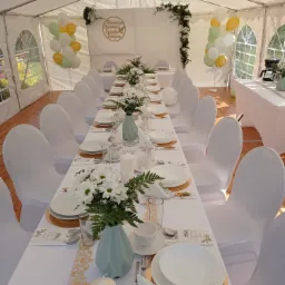 Uroczysty stół komunijny w białym namiocie: białe krzesła z pokrowcami, zastawa na złotych podkładkach, wazoniki z białymi kwiatami, balony w kolorach bieli, zieleni i złota, dekoracja z zieleni...