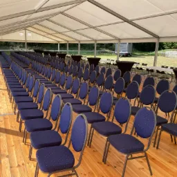 Wnętrze białego namiotu z rzędami krzeseł w kolorze granatowym w białe kropki, ustawionych na drewnianej podłodze, w tle stoliki koktajlowe z czarnymi obrusami i widok na zieleń za namiotem.