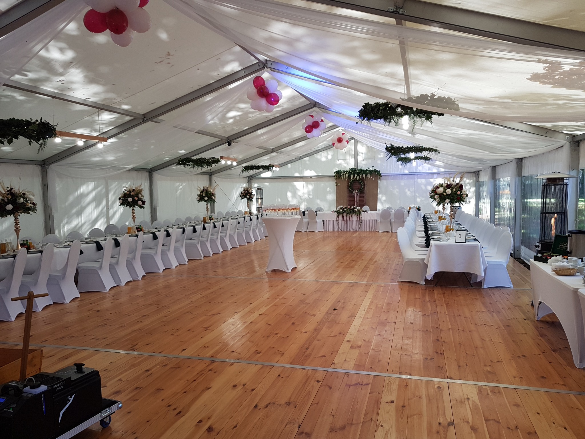 Wnętrze białego namiotu bankietowego z drewnianą podłogą, udekorowanego kwiatami, girlandami i balonami, przygotowanego na uroczystość.