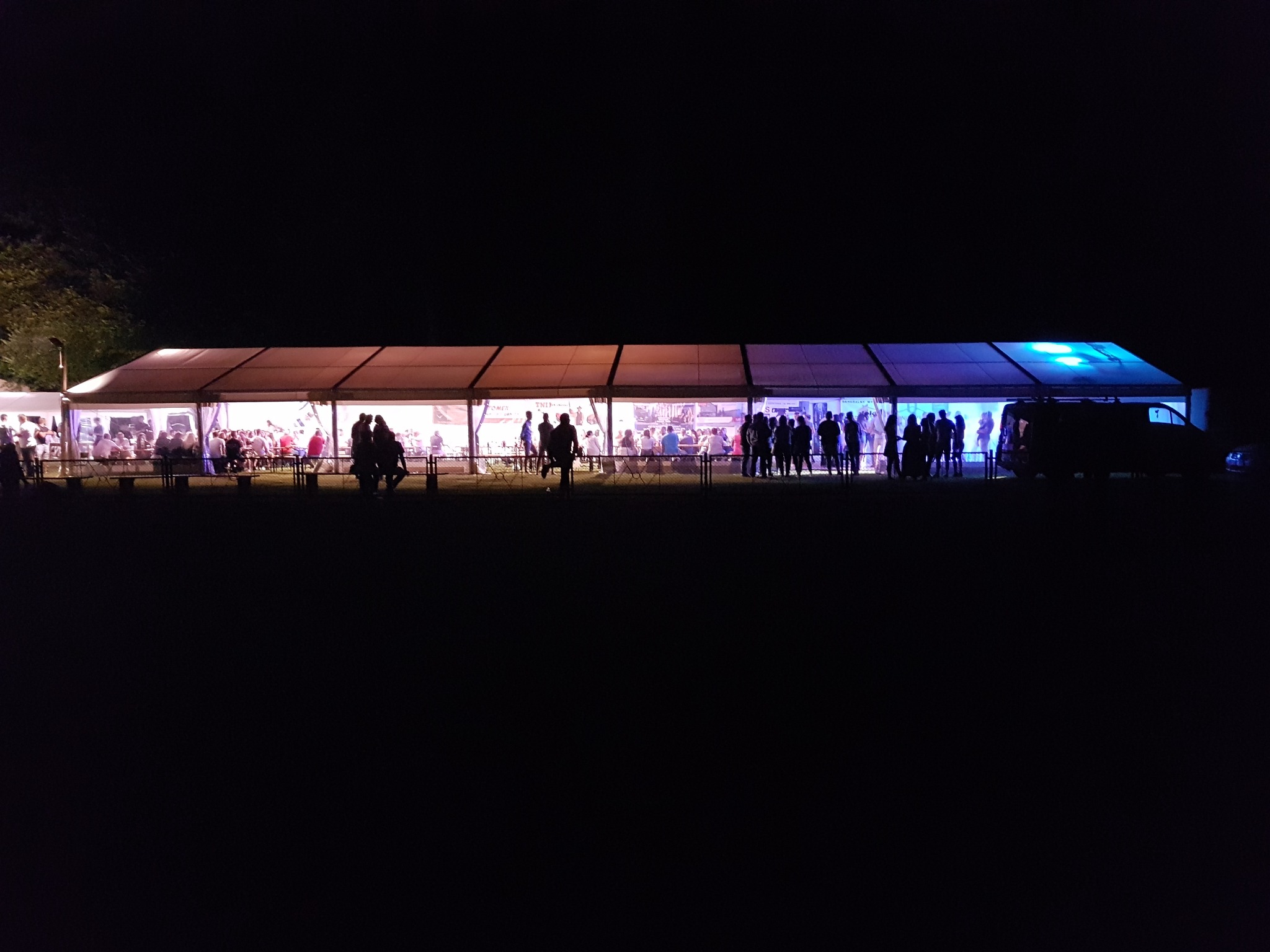 Oświetlony namiot imprezowy nocą, widoczne sylwetki ludzi wewnątrz i na zewnątrz, oświetlenie w kolorach pomarańczowym i niebieskim, ciemne otoczenie.