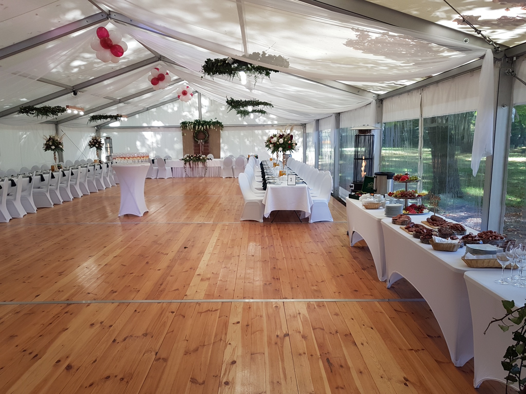 Uroczysty namiot z drewnianą podłogą, dekoracjami kwiatowymi, balonami i stołami z białymi obrusami, przygotowany do przyjęcia gości, widoczny stół z przekąskami.