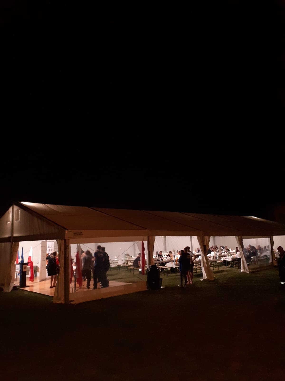 Oświetlony namiot eventowy z gośćmi wewnątrz, widoczne flagi Polski i Unii Europejskiej przy wejściu, wieczorne oświetlenie.