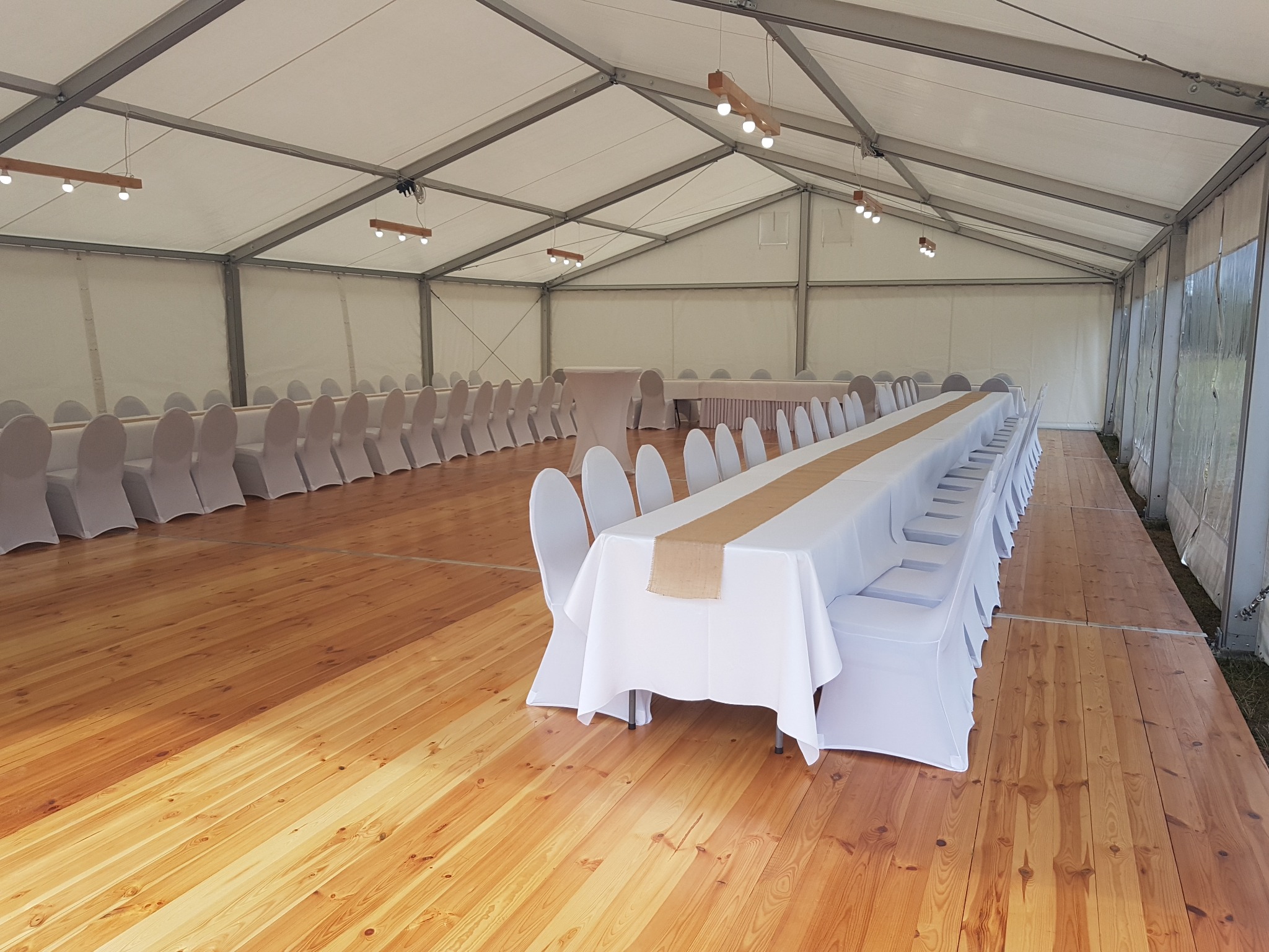 Wnętrze białego namiotu bankietowego z drewnianą podłogą, długie stoły z białymi obrusami i jutowymi bieżnikami, krzesła w białych pokrowcach, oświetlenie w postaci drewnianych belek z żarówkami.