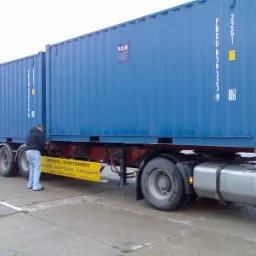 Ciężarówka z dwoma niebieskimi kontenerami, mężczyzna w bluzie i jeansach sprawdza mocowanie ładunku, widoczny żółty baner reklamowy firmy transportowej.