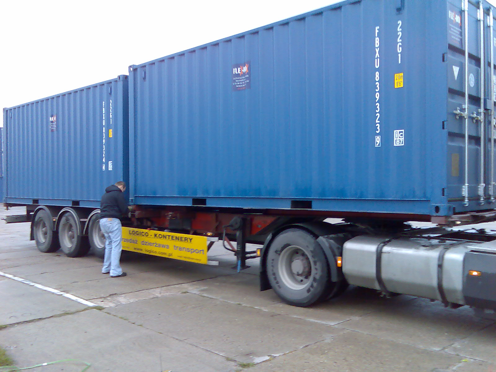 Ciężarówka z dwoma niebieskimi kontenerami, mężczyzna w bluzie i jeansach sprawdza mocowanie ładunku, widoczny żółty baner reklamowy firmy transportowej.