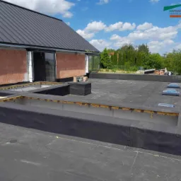 Widok z góry na płaski dach pokryty czarną membraną, z widocznymi świetlikami i obróbkami attyki, budynek w trakcie budowy z czerwoną cegłą i oknami przesuwnymi, logo firmy w prawym górnym rogu.