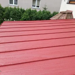 Czerwony dach z blachy trapezowej po deszczu, widoczne krople wody, w tle drabina oparta o ścianę budynku i zielone tuje.