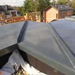 Szary dach z blachy na budynku w trakcie budowy, widoczne obróbki blacharskie, zabezpieczony białą tkaniną, w tle budynek z cegły i konstrukcja drewniana.