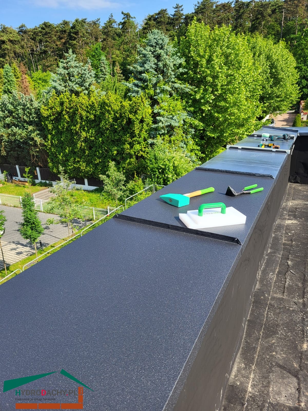 Czarny, płaski dach po renowacji w Myślenicach. Na dachu leżą narzędzia dekarskie: młotek, szpachelka i inne. W tle zieleń drzew i krzewów.