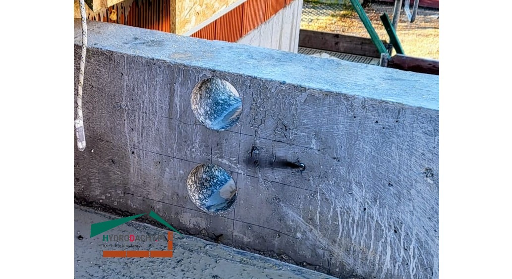 Betonowy element konstrukcyjny z dwoma wywierconymi otworami, widoczne ślady po wierceniu, sznur i logo firmy w rogu.