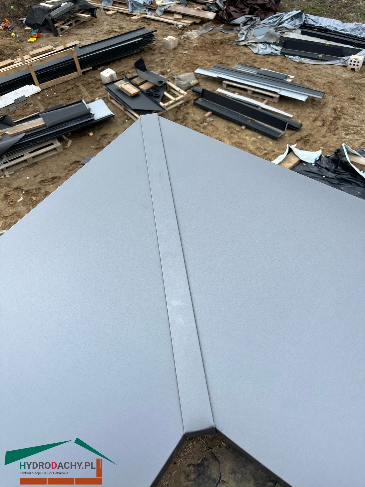 Szary, metalowy dach z widocznym łączeniem paneli na tle placu budowy z elementami konstrukcji dachowej i logo firmy dekarskiej w rogu.