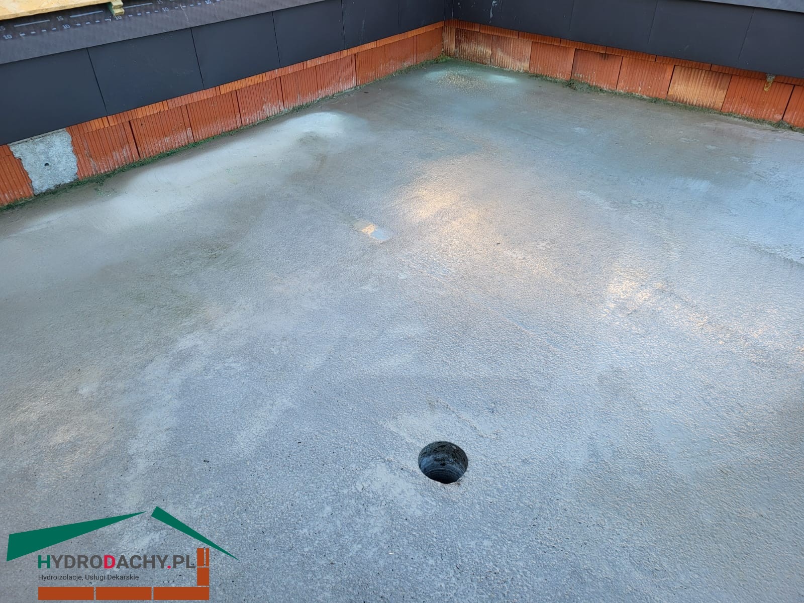 Świeżo wylana, mokra wylewka na dachu z widocznym odpływem i fragmentem muru. W tle czarna, pionowa powierzchnia. Logo firmy w rogu kadru.
