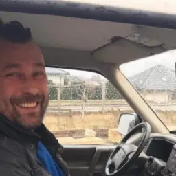 Uśmiechnięty mężczyzna z irokezem siedzi za kierownicą samochodu dostawczego, widok na domy w tle przez szybę.